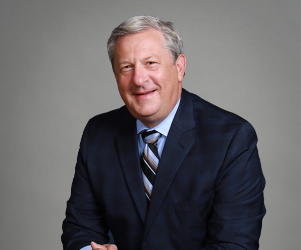 A professional portrait of Adam Zucker, a smiling man in a dark blue suit with a plaid pattern and a light blue tie, seated against a neutral gray background.