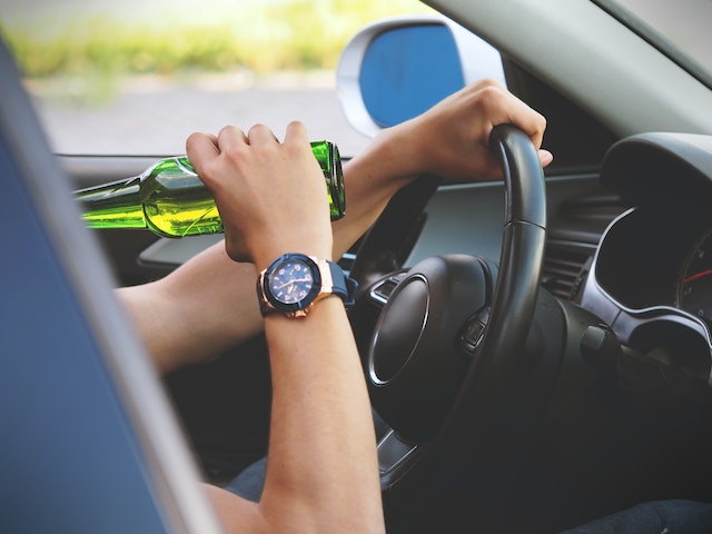 A person driving a car while holding a green bottle of beer in Pennsylvania, implying unsafe driving behavior. Their hands are on the steering wheel, wearing a watch.