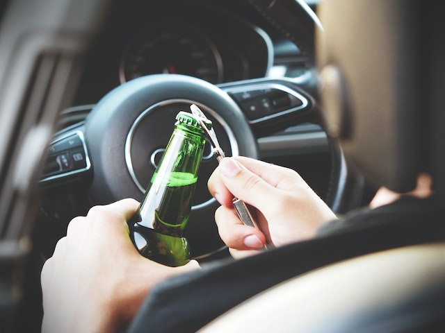 A person holding a beer bottle with a cap opener while sitting in the driver's seat of a car in Pennsylvania, depicting an irresponsible or illegal act of drinking and driving.